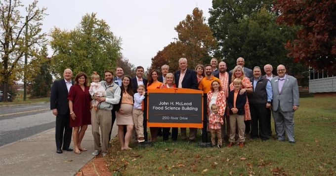 A group of people of various ages gathered around a sign that reads "John H. McLeod Food Science Building, 2520 River Drive," in front of a tree-lined background.