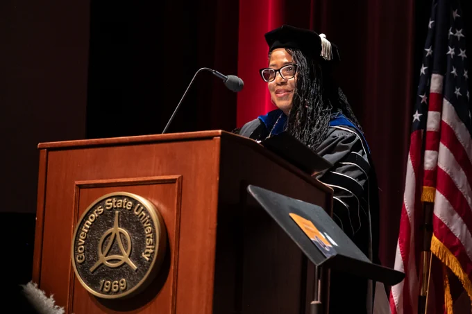 GovState President in academic regalia speaking at a podium with the Governors State University seal and an American flag in the background.