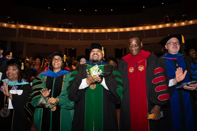 Graduates in academic regalia clapping joyfully at a investiture ceremony in a theater.