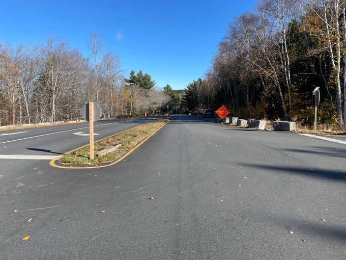 Empty paved road with a small traffic island and autumn trees under a clear blue sky.