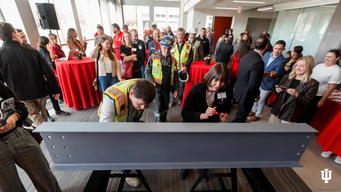 Attendees at an Indiana University event are gathered around a large sign being signed by two individuals, with a festive atmosphere indicated by red-covered tables and casual conversations.