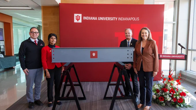Four individuals standing in front of a red Indiana University Indianapolis backdrop, with floral arrangements at the base.