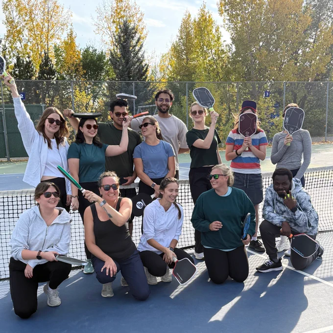 The ENHANCE lab team in a group photo on a pickleball court, laughing together with their racquets raised, after learning to play pickleball. 