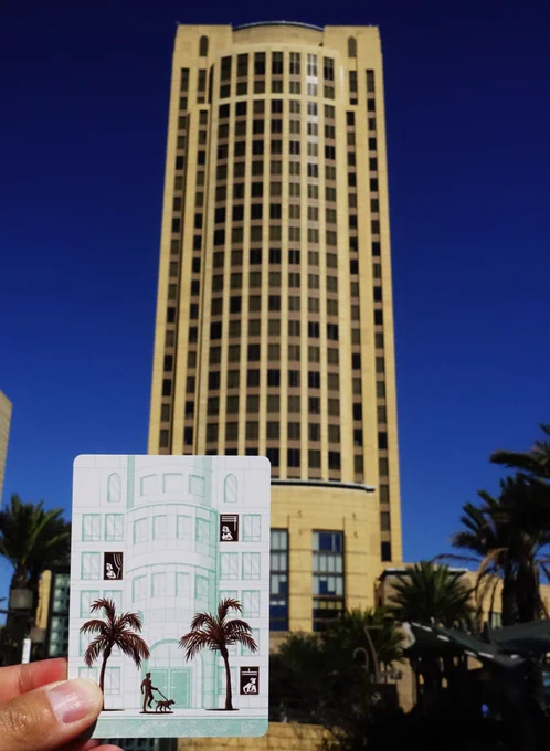 A card from the card game Downtown Las Palmas is held in front of the Metro Headquarters Building in Downtown Los Angeles. The drawing on the card was inspired by the building.