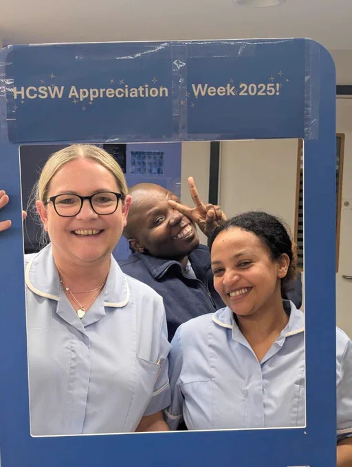 Three healthcare staff members wearing light blue uniforms pose indoors while holding a large blue photo frame. The frame has white text at the top reading “HCSW Appreciation Week 2025!” and features the Greater Manchester Mental Health NHS Foundation Trust logo. The background shows an indoor setting with neutral walls and a door partially visible.