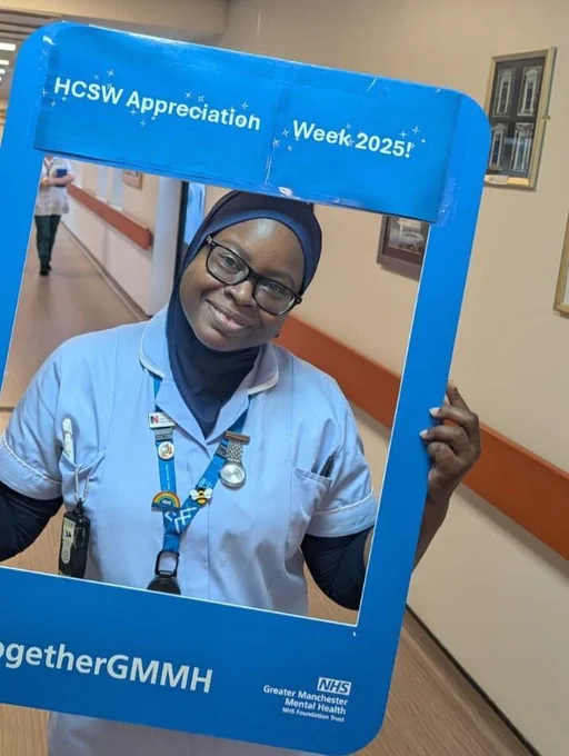 A healthcare colleague wearing a light blue uniform and a lanyard with multiple badges is standing in a hospital corridor, holding a large blue photo frame. The frame has white text at the top reading “HCSW Appreciation Week 2025!” and at the bottom “#TogetherGMMH” along with the Greater Manchester Mental Health NHS Foundation Trust logo. The corridor has cream-colored walls with orange accents and framed pictures on the wall.