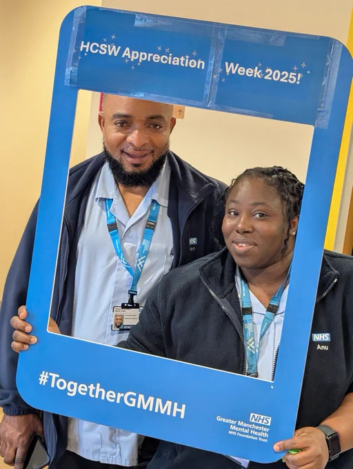 Two healthcare colleagues hold a large blue photo frame with white text that reads “HCSW Appreciation Week 2025!” at the top and “#TogetherGMMH” at the bottom. The frame also includes the Greater Manchester Mental Health NHS Foundation Trust logo. The background shows an indoor setting with neutral-coloured walls.