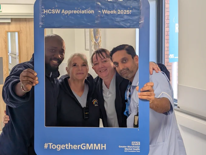 Four healthcare colleagues standing close together indoors, holding a large blue photo frame with white text that reads “HCSW Appreciation Week 2025!” at the top and “#TogetherGMMH” at the bottom. The frame also includes the Greater Manchester Mental Health NHS Foundation Trust logo. The background includes a wooden door on the left and a bright window on the right.