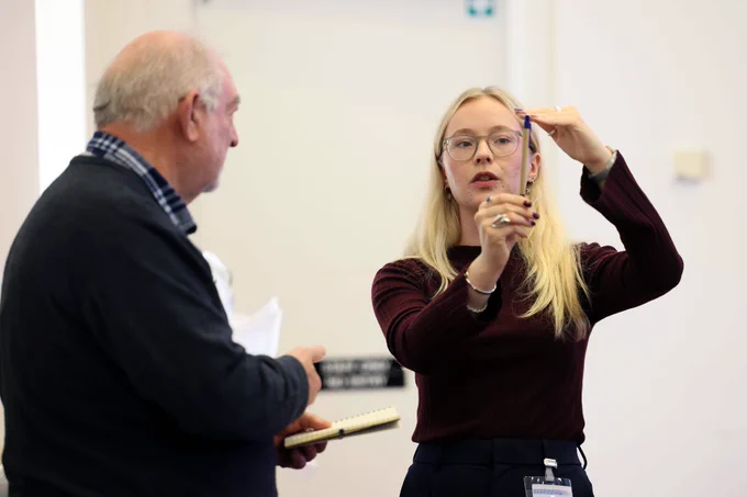 A lady with blonde hair and a maroon jumper talking to a man in a dark jumper with a checked collar. 