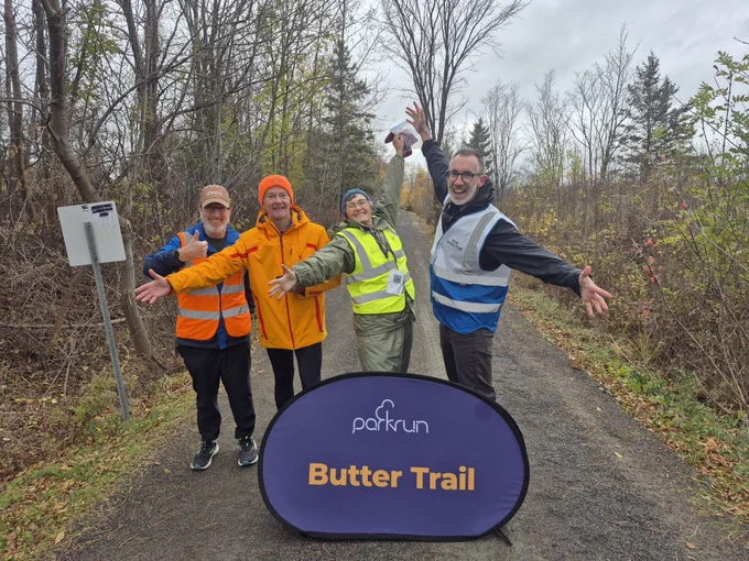 Four parkrunners posing joyfully behind the 'Butter Trail' parkrun purpil panel, with autumn trees in the background.