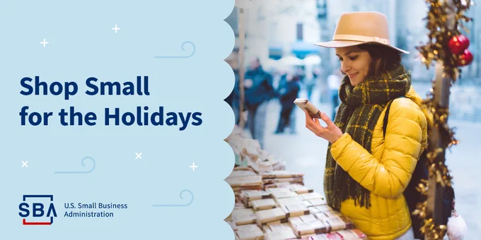 Promotional image from the U.S. Small Business Administration encouraging shopping at small businesses during the holidays. Features an individual in a yellow scarf and hat browsing on a smartphone while standing by a market stall.