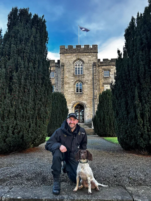 PC Connell kneels beside a brown and white detection dog, PD Anna. Behind them is Tulliallan castle with a Scottish flag flying from the top.