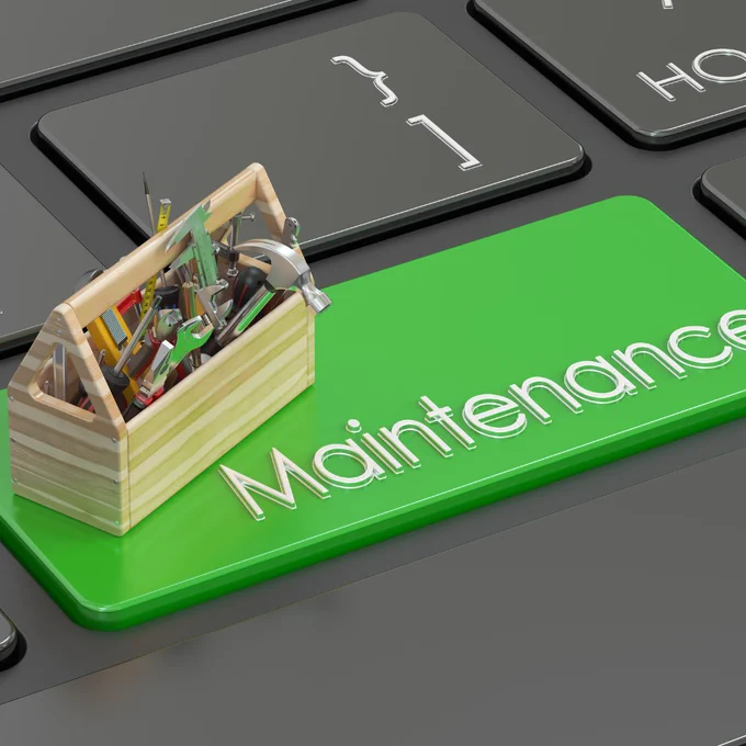 A close-up of a computer keyboard with one large green key labelled “Maintenance.” On top of the key sits a small wooden toolbox filled with assorted tools, including a hammer, wrench, screwdriver, tape measure, and pliers. The surrounding keys are black, making the bright green maintenance key stand out.