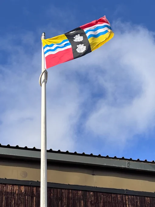 The Bedfordshire flag raised above Biggleswade rugby club.
The flag's yellow and red quadrants are taken from the arms of the Beauchamp family, powerful in the county after the Norman conquest of England and constructors of Bedford Castle. The blue and white wavy lines symbolise the Great Ouse, which flows through the county. The three shells are taken from the coat of arms of the Duke of Bedford.