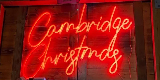 Neon sign reading "Cambridge Christmas" illuminated in red, displayed at a festive outdoor setting with decorative lights and greenery.