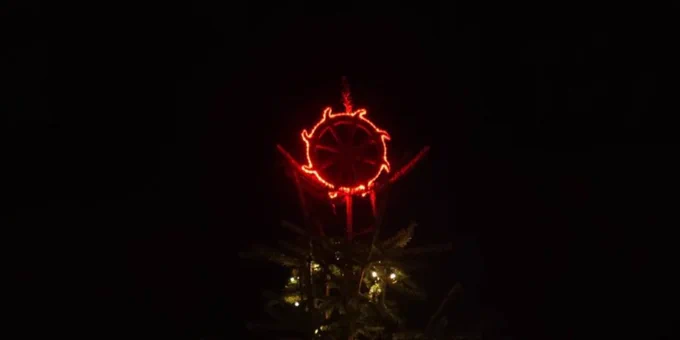 A Christmas tree illuminated with twinkling lights, topped with a unique red Catharine Wheel decoration, framed by an archway in the foreground, with dimly lit buildings visible in the background.