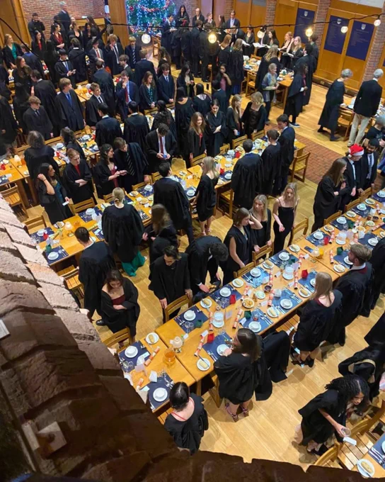 A bird's eye view of Robinson College's grand dining hall. Rows of long tables topped with festive table settings. College members in formal gowns are standing by their places. A Christmas tree is at the back.