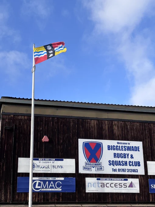 The Bedfordshire flag raised above Biggleswade rugby club.
The flag's yellow and red quadrants are taken from the arms of the Beauchamp family, powerful in the county after the Norman conquest of England and constructors of Bedford Castle. The blue and white wavy lines symbolise the Great Ouse, which flows through the county. The three shells are taken from the coat of arms of the Duke of Bedford.