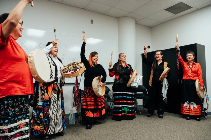 Six members of the Khast'an Drummers performing a traditional drum dance in a room, wearing colourful ribbon skirts.