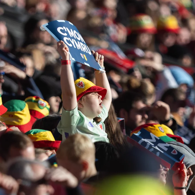 A girl in the stadium stands holding a ‘C’mon Cymru’ sign, cheering enthusiastically for the Wales football team.