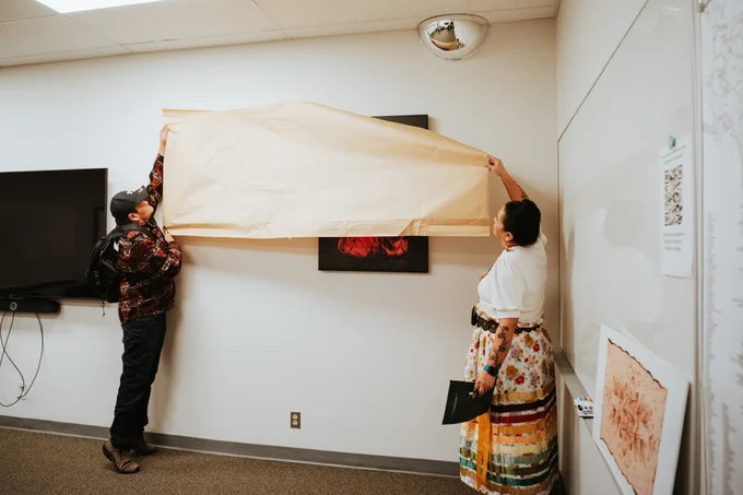 Two individuals unveil a plaque and portrait in a room with a display screen and artistic elements on the walls.