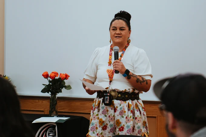 Person giving a speech in front of an audience, wearing a floral skirt, white blouse and lanyard and holding a microphone.