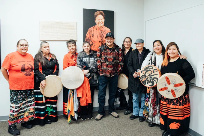 Group of individuals holding traditional drums standing in front of a portrait in an indoor setting.