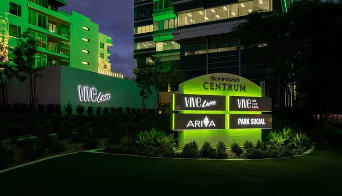 Illuminated modern building signage at night displaying business names surrounded by greenery in an urban commercial area.