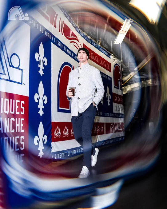 Brock Nelson during arrivals with a cold brew coffee in hand wearing blue dress pants, a grey button up jacket, a black beanie and white sneakers. The background features Nordiques logos and imagery