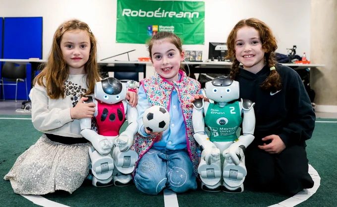 Three young girls kneel on a green floor, each posing with a small humanoid robot. The girl on the left wears a cream top, the middle one in a colorful jacket holds a soccer ball, and the right girl is in a black outfit. The robots, dressed in pink and green jerseys, sit between them. A "RoboÉireann" banner is visible in the background.