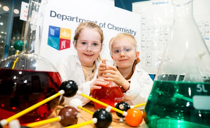 Two children wearing safety goggles and lab coats smile while holding a beaker with red liquid. They are in a chemistry lab setting, surrounded by large flasks containing colorful liquids and molecular models. Behind them is a periodic table and a sign reading "Department of Chemistry," creating a vibrant, educational atmosphere.