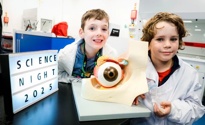 Two children in lab coats are smiling at the camera while displaying a large anatomical model of an eye. Beside them is a lightbox sign reading "Science Night 2025." The background shows a modern laboratory setting with blue cabinets.