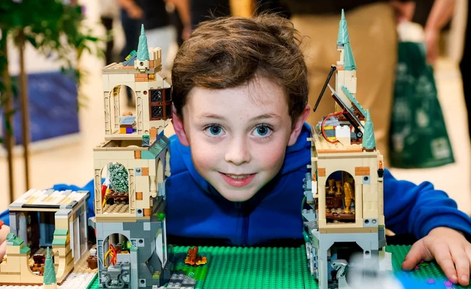 A young child in a blue hoodie smiles while playing with a detailed, multicolored LEGO castle set, peering through the gaps in the structure. The castle features intricate towers, arches, and architectural details with green and beige bricks. The scene exudes creativity and fun, with out-of-focus people and plants in the background.