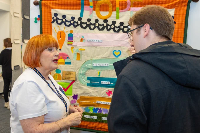 An event participant sharing the story behind the Peer Support Hope Quilt with another participant. Both are standing in front of the quilt.