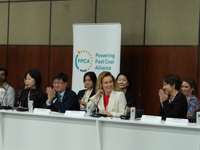 A group of individuals seated at a long table during a conference or meeting, with a banner in the background that reads "PPCA: Powering Past Coal Alliance." 