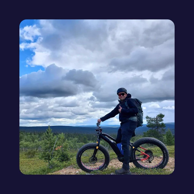 Jukka-Pekka wearing dark outdoor gear and sunglasses is standing with a fatbike on a forested hilltop. He’s giving a thumbs-up while looking at the camera. The sky is cloudy with patches of blue, and pine trees stretch into the distance below.