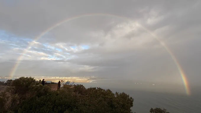 A full, bright rainbow arches across a cloudy sky over San Diego Bay. Sunlight breaks through the clouds, lighting up the coastline and the ocean below. A few visitors stand near the overlook, enjoying the view while coastal shrubs frame the foreground. 