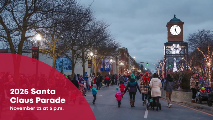 A photo of people walking up a closed street. Holiday lights are strung through the trees and people are dressed for cold weather. Text on the photo reads, "2025 Santa Claus Parade. November 22 at 5 p.m."