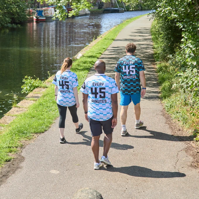 Three people wearing Prostate United shirts with 'EVERY 45 MINUTES' written on them are walking along a canal path, surrounded by lush greenery.