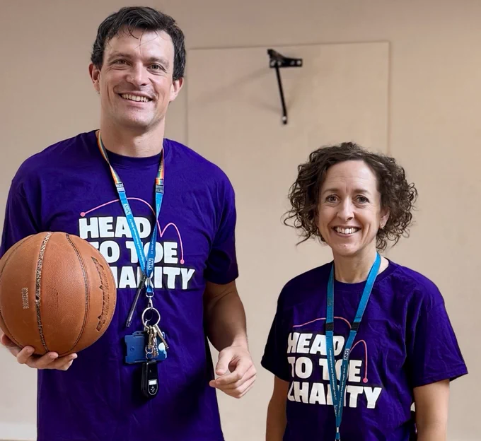 Two individuals wearing "Head to toe Charity" t-shirts, smiling while holding a basketball, standing indoors.