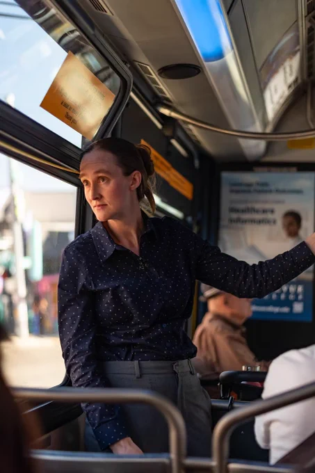 an image of Katie Wilson standing in a button-up shirt and slacks riding the bus in the morning while holding on onto a rail

