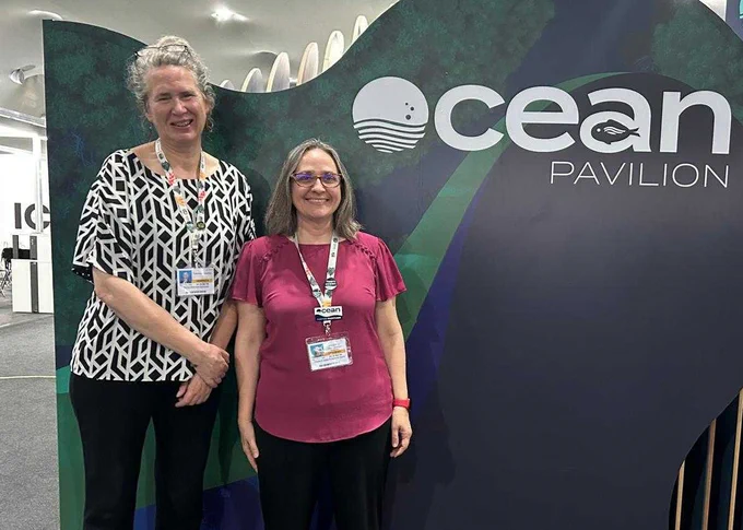 Drs. Sonya Dyhrman and Elizabeth Kujawinski stand together in front of the Ocean Pavilion sign during COP30 in Brazil. Photo provided by Elizabeth Kujawinski. 