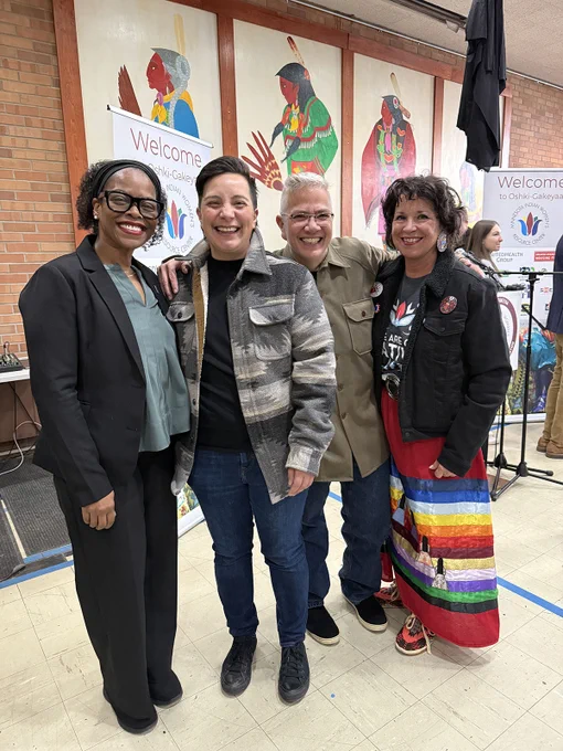 Four adults smiling at a community event, standing in front of a "Welcome" banner and colorful artwork. Two are dressed in business casual clothes, and one is wearing a multi-colored ribbon skirt.