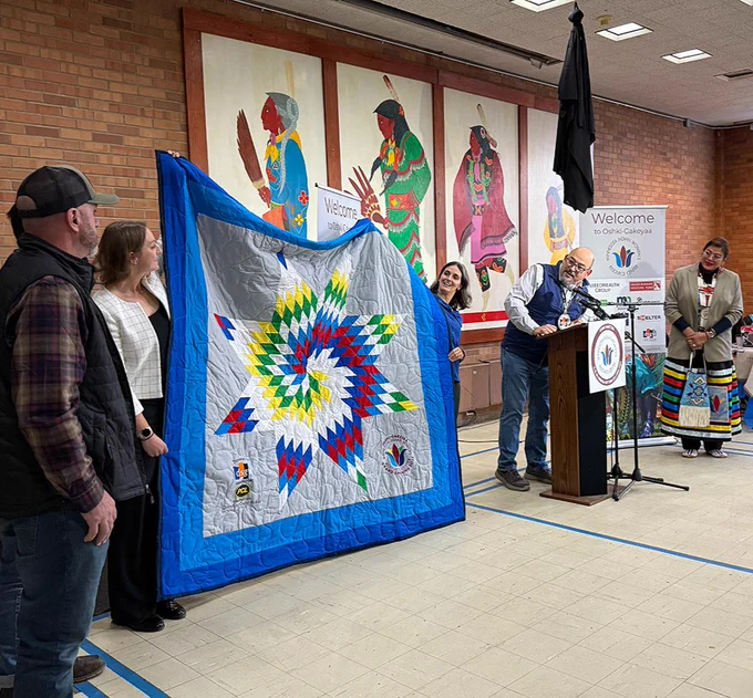 A group of individuals at a public event, displaying a large, colorful quilt with starburst patterns. A speaker stands at a podium with microphones, and banners reading "Welcome" are displayed in the background, along with vibrant Native artwork.