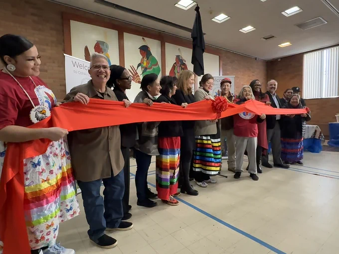 A dozen individuals hold a red ribbon at a ceremonial ribbon-cutting event, with Native art in the background.