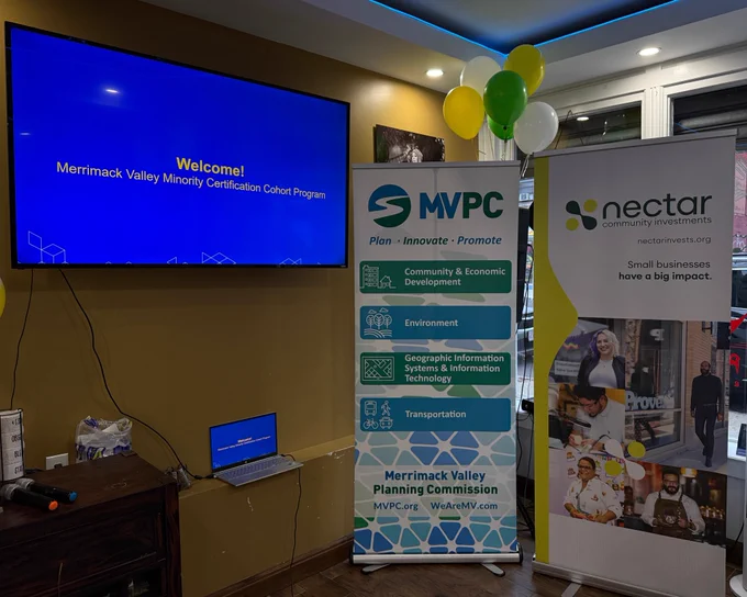 A photo of the entrance to the event. A TV has yellow text on a blue background and reads "Welcome! Merrimack Valley Minority Certification Cohort Program." A banner highlights the Merrimack Valley Planning Commission. A second banner is for Nectar Community Investments.