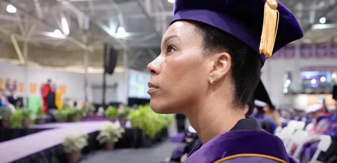 Michelle Lollie in profile, her attention directed to commencement ceremony proceedings. Her expression is resolute. Michelle wears her purple and gold graduation regalia. 