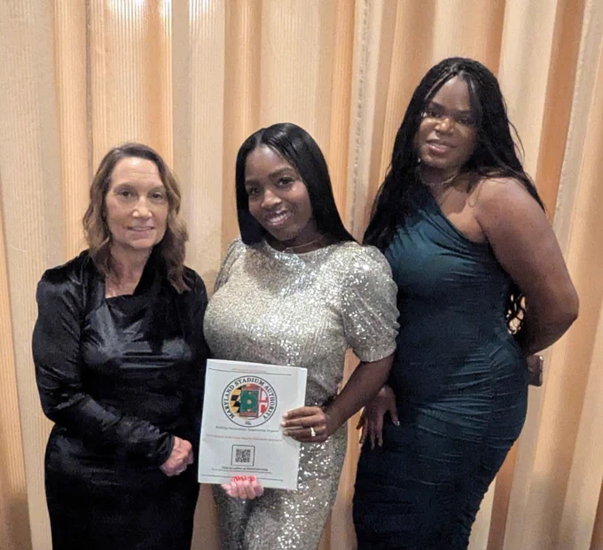 Three people posing for a photo at an  at the recent Maryland Minority Contractors Association 12th Soirée & Awards Banquet​, dressed in formal attire with one holding a advertisement featuring the emblem of the Maryland Stadium Authority and QR code.