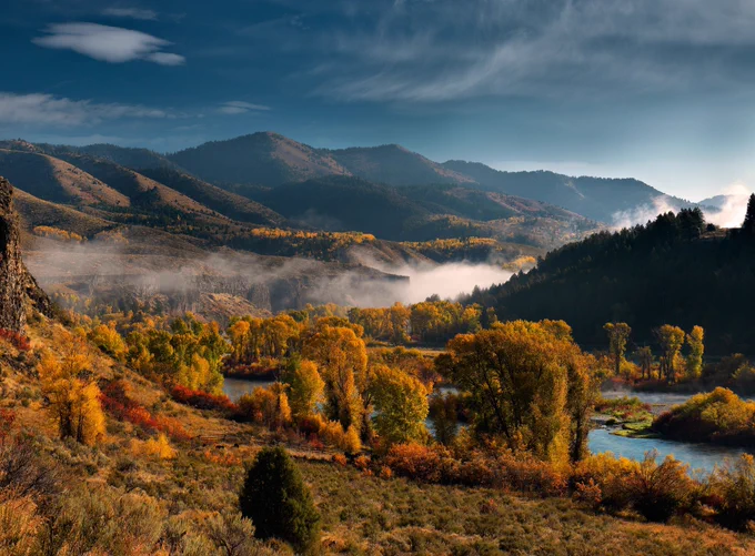 Autumn landscape with colorful trees, a winding river, and mist rising in a valley surrounded by mountains under a partly cloudy sky.