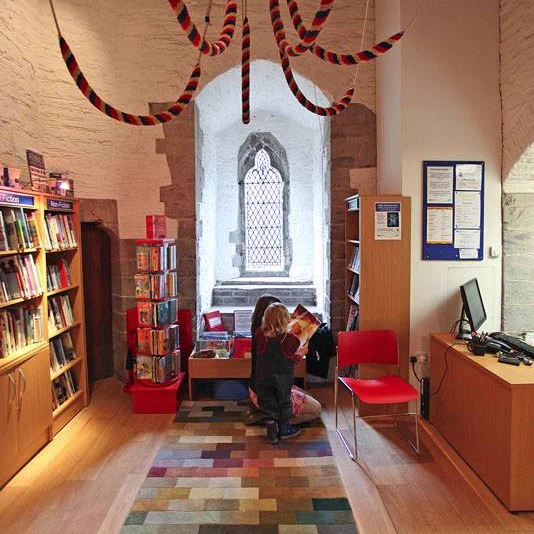 Internal view of Peterchurch Library. The room has high ceilings and the walls are white washed stone blocks. The room is lined with book shelves and illuminated by a vaulted, leaded window.   A woman and child kneel on a rug reading a picture book together.